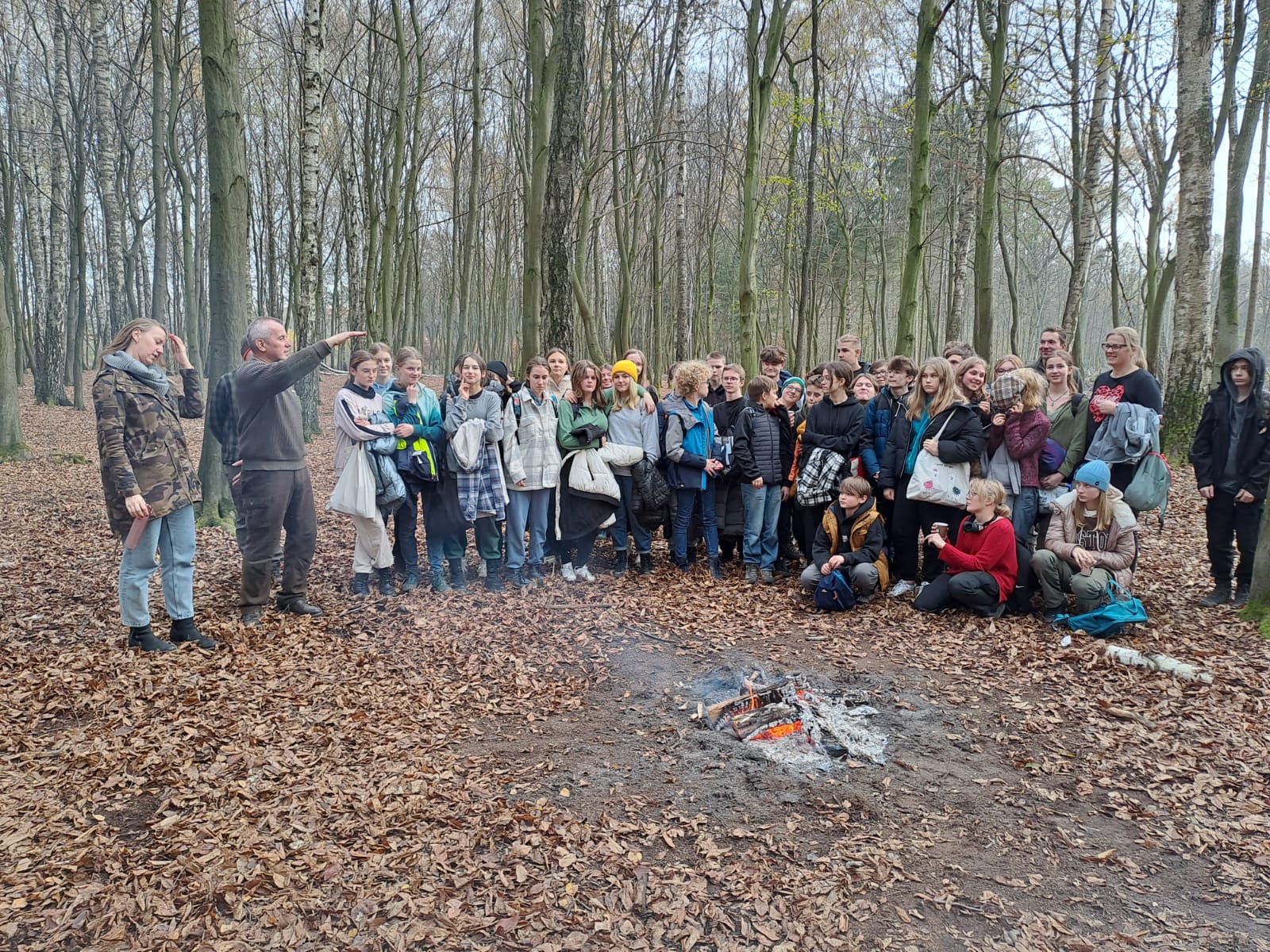 Zdjęcie na tle lasu przedstawia grupę uczniów przy ognisku oraz leśniczego prowadzącego edukację leśną. Fot. Krzysztof Radke 
